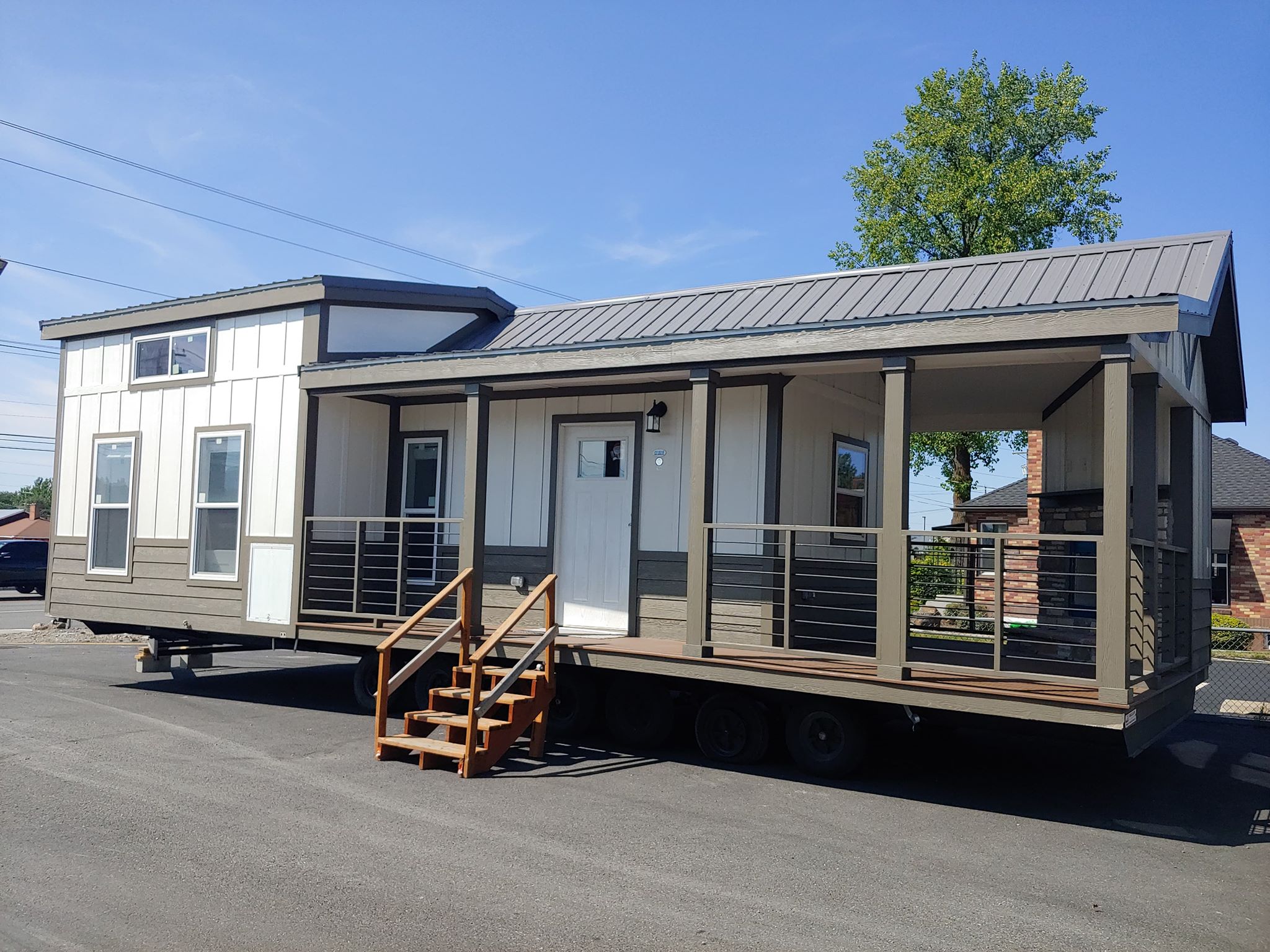 A modern tiny house with a pitched metal roof and large windows, set on wheels in a parking lot. It has a porch with railings and wooden steps.
