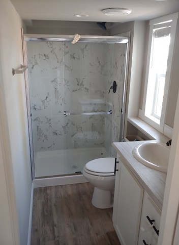 Small bathroom with a glass-enclosed shower featuring a marbled tile design. Wooden floor, white vanity with a round sink, and two narrow windows. Bright and clean.