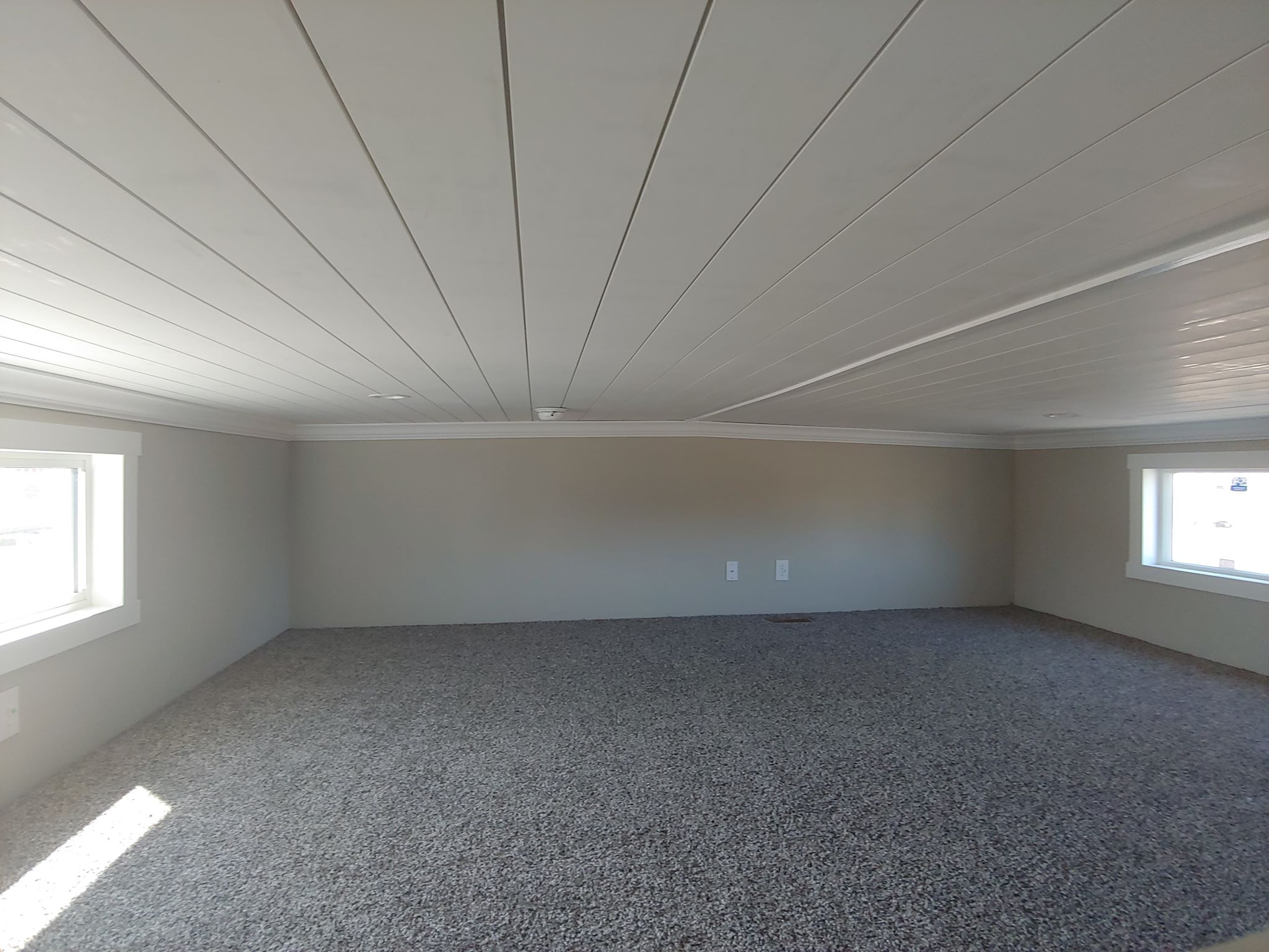 A low-ceilinged room with beige walls and a gray carpeted floor. Two small windows on opposite walls allow natural light to enter, creating a cozy atmosphere.
