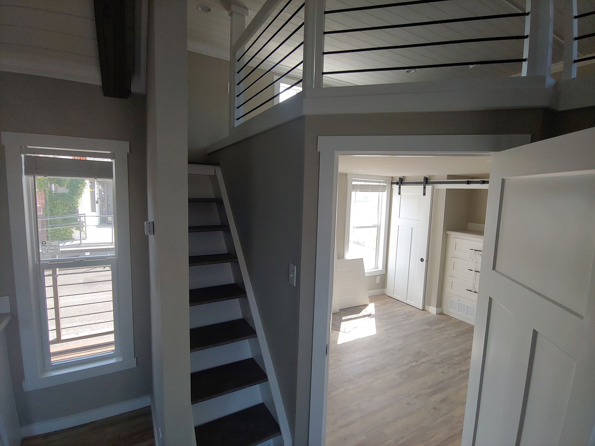A modern, small interior with a narrow staircase leading to a loft. Sunlight streams through large windows in a room with light wood flooring and sliding door.