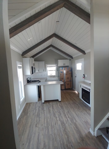 Compact, rustic kitchen with vaulted ceiling and wooden beams. Features white cabinets, a small island, boxes by the fridge, and wood-style flooring.
