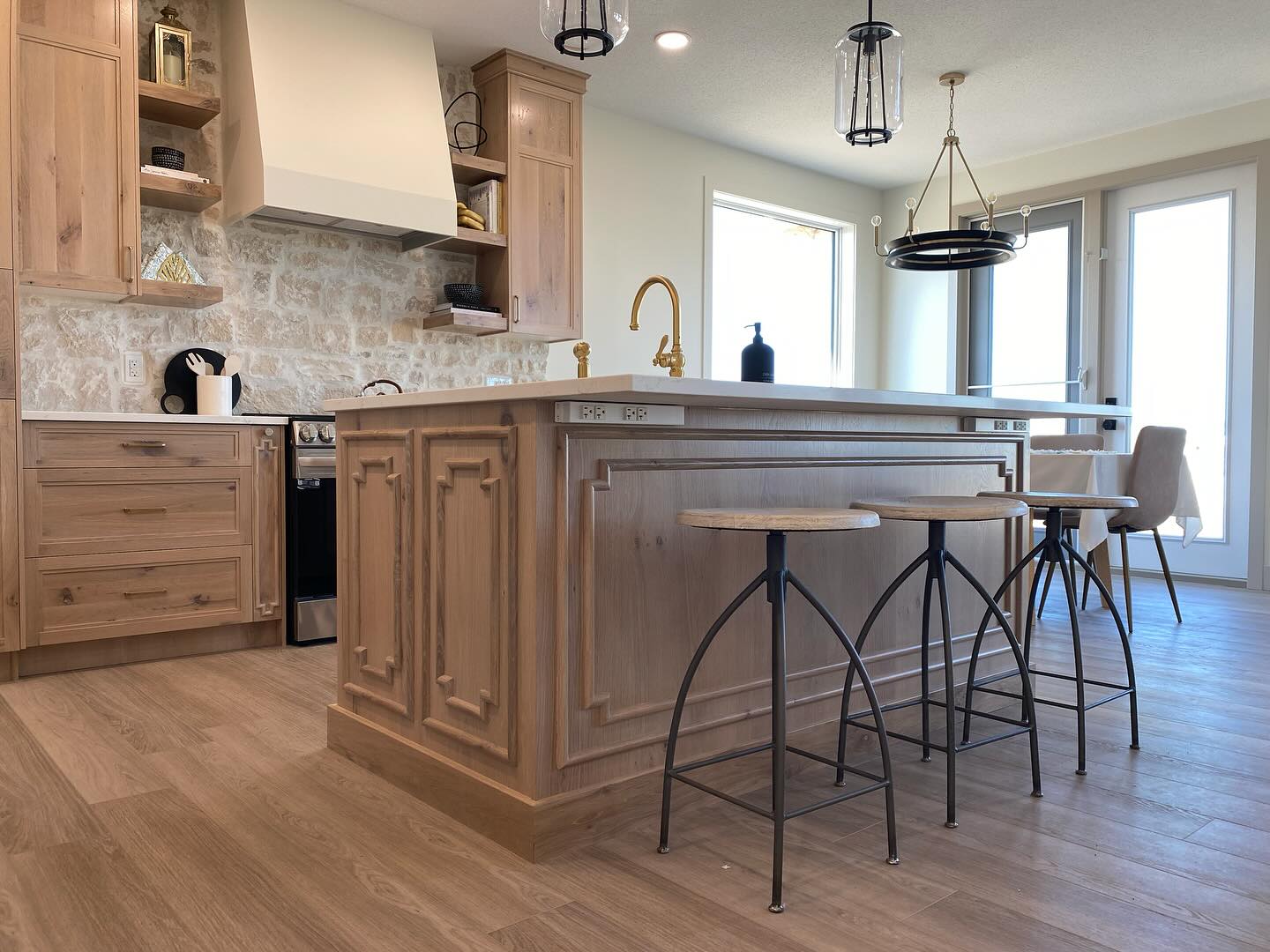 Warm, rustic kitchen with wooden cabinetry and island, stone backsplash, gold faucet, bar stools, pendant lights, and sunlight from large windows.