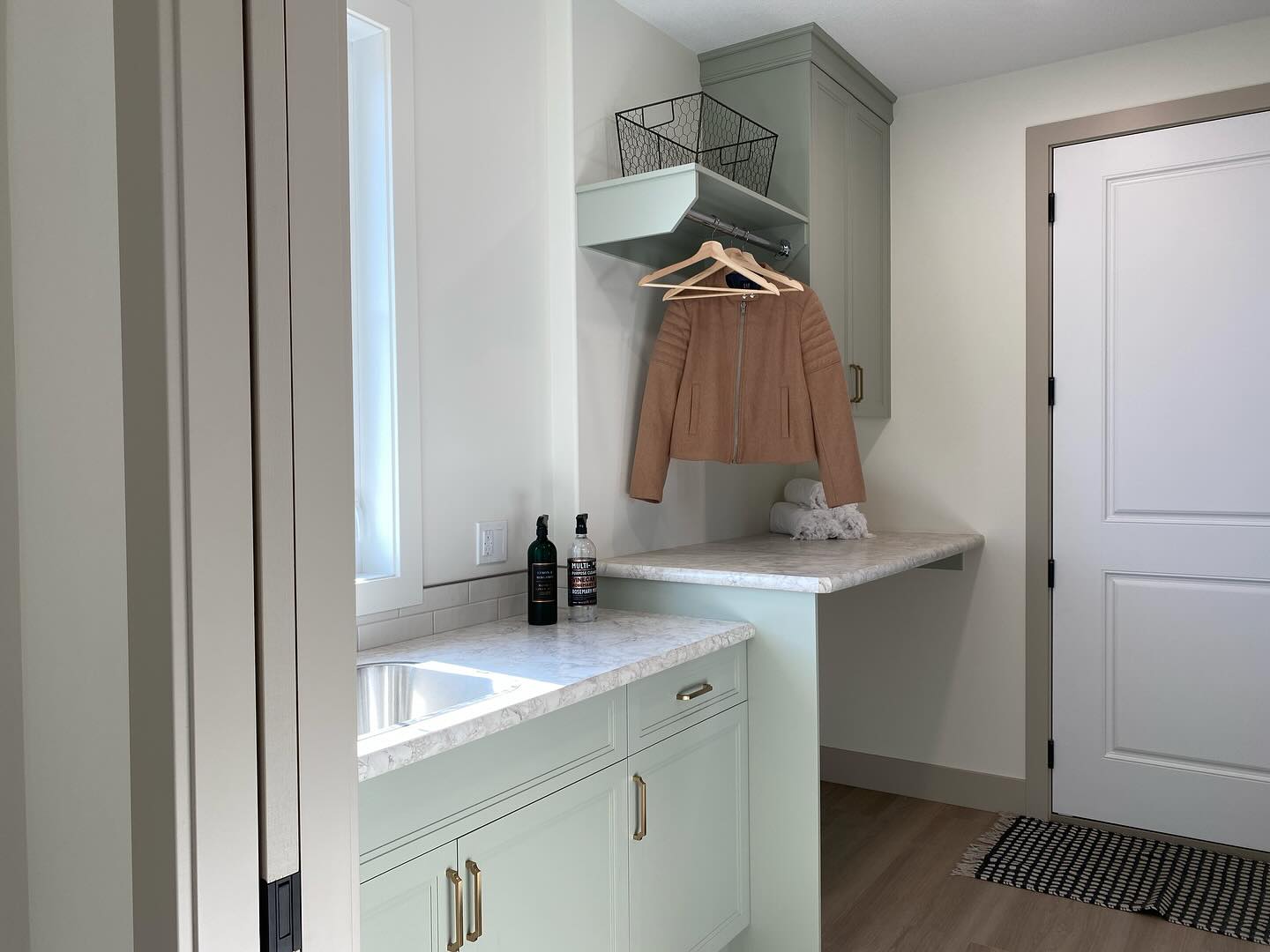 A tidy laundry room with mint green cabinets, a marble countertop, and a sink. A tan jacket hangs above a folded towel, creating a fresh and organized atmosphere.
