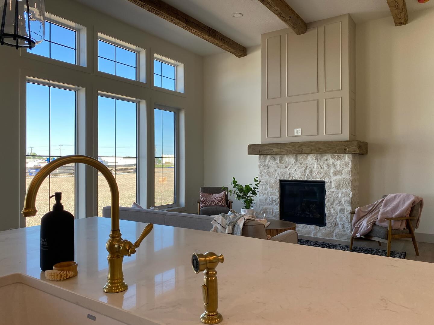 Spacious living room with large windows, wooden beams, and stone fireplace. Cozy seating area with soft lighting. Elegant brass faucet visible in foreground.