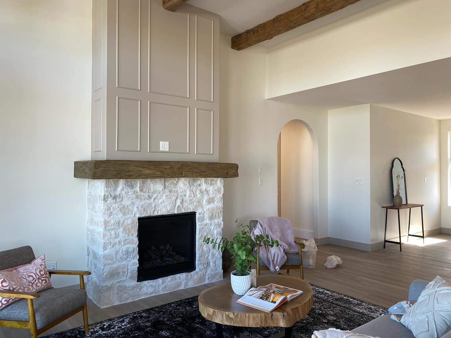 Cozy living room with a stone fireplace and wooden mantel. Armchairs and a round coffee table with a plant add warmth. Arched doorway and soft lighting enhance the inviting atmosphere.