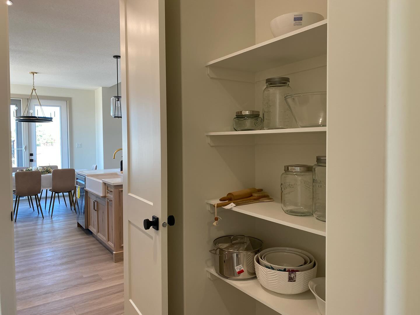 Pantry with neatly arranged jars and bowls on shelves, next to a modern kitchen and dining area with wooden floors and natural light.