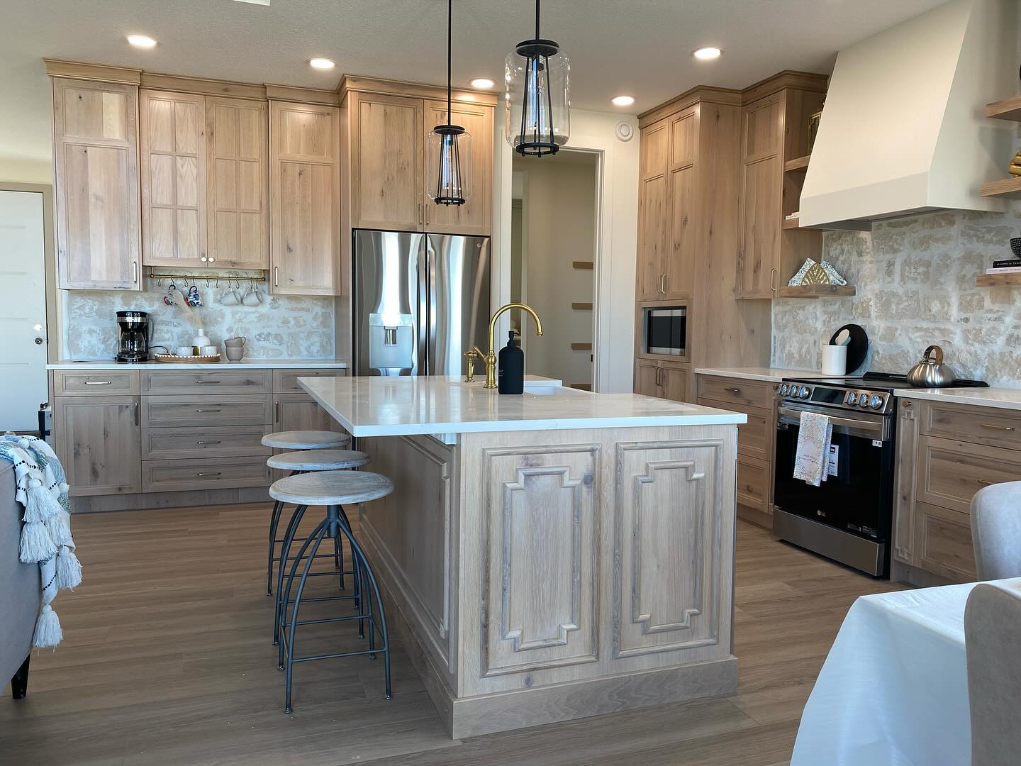 A modern kitchen with light wood cabinets, a large island with bar stools, stainless steel appliances, and stone backsplash. Pendant lights add elegance.