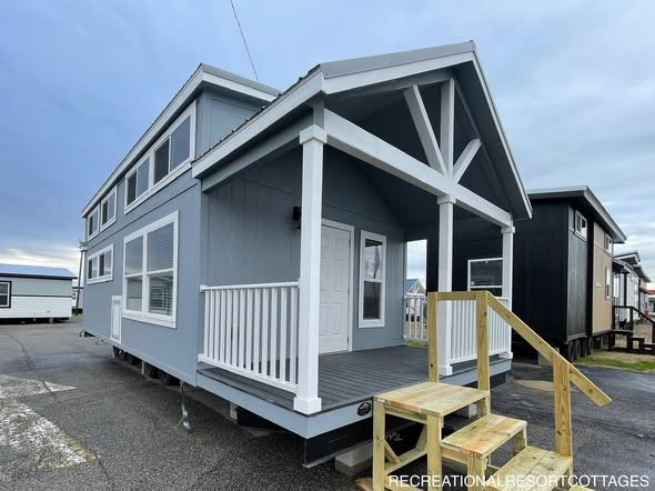 A modern tiny house with a blue exterior, white trim, and gabled roof. Front porch with white railing and wooden steps. Overcast sky in the background.