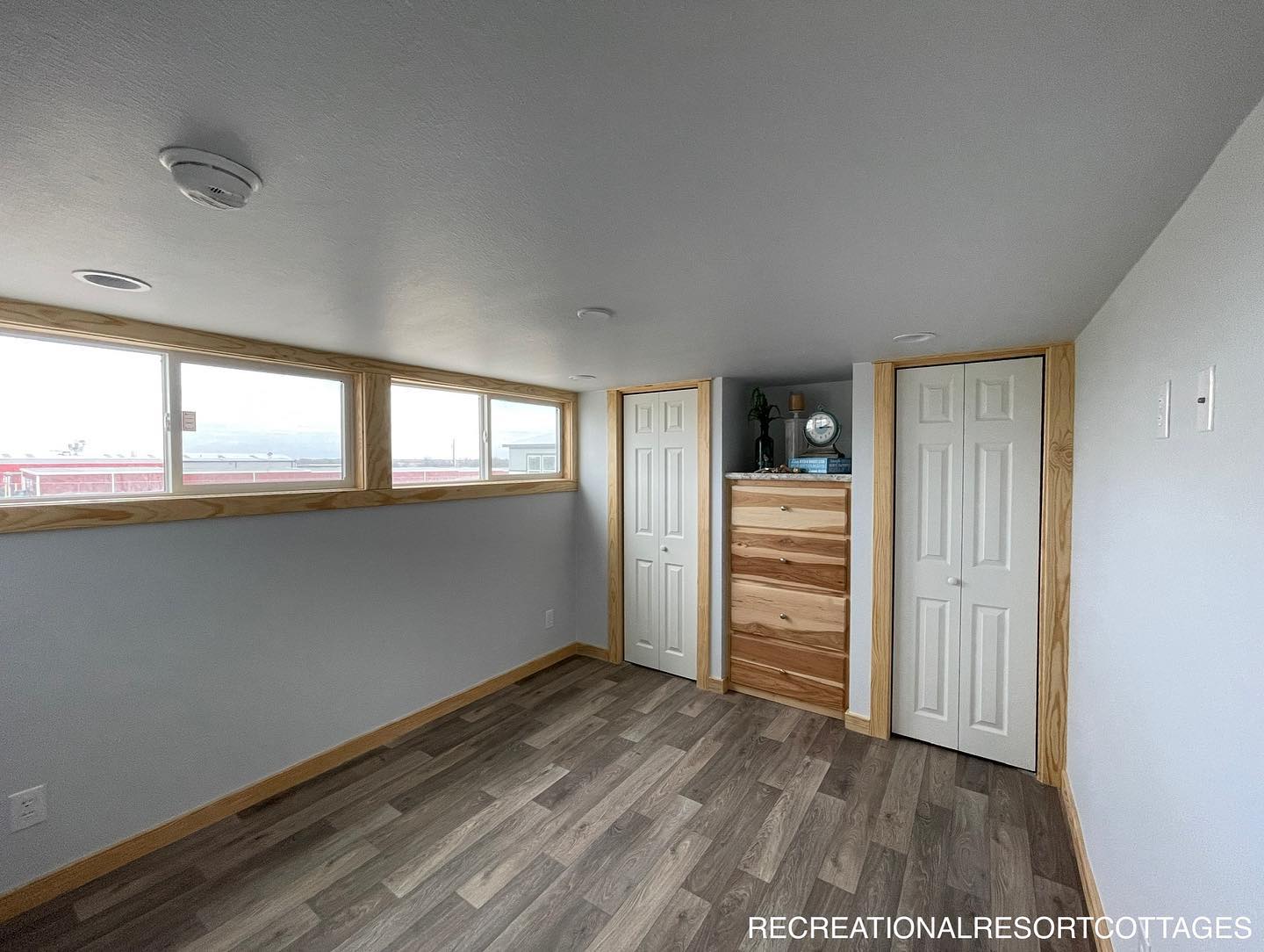 A small room with light wood flooring and white walls. It features three upper windows, two closets with white doors, and a wooden dresser. Bright and minimalist.