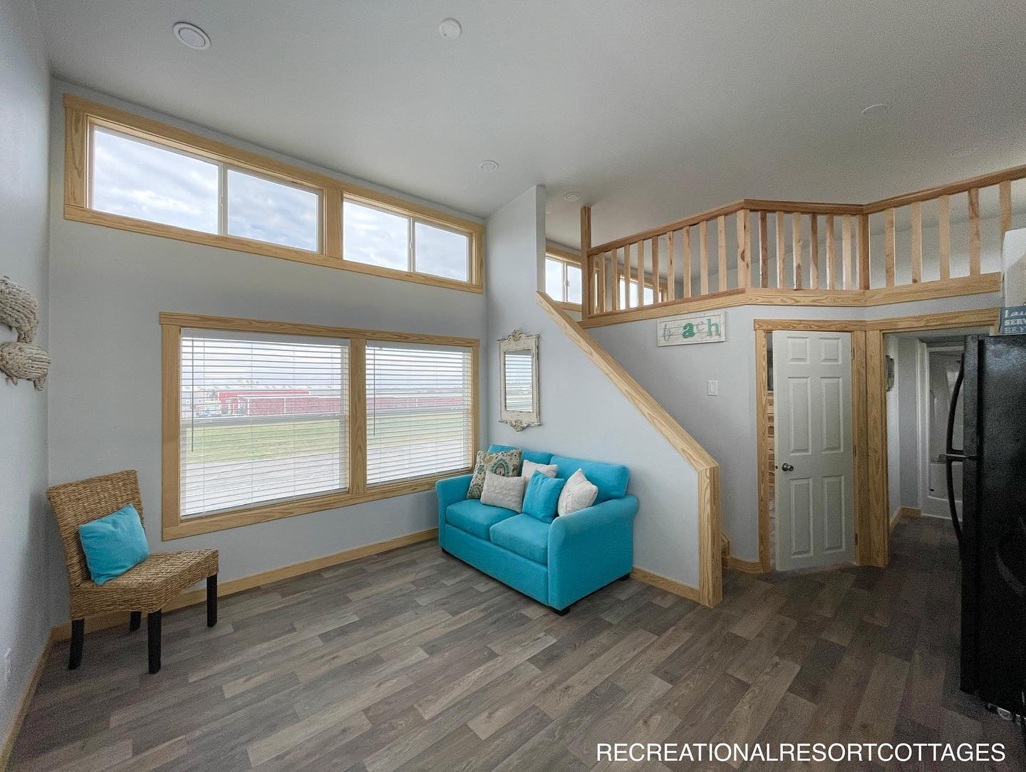 Bright living room with large windows, a turquoise sofa, and a wicker chair. A staircase leads to a loft. Light gray walls create an inviting atmosphere.