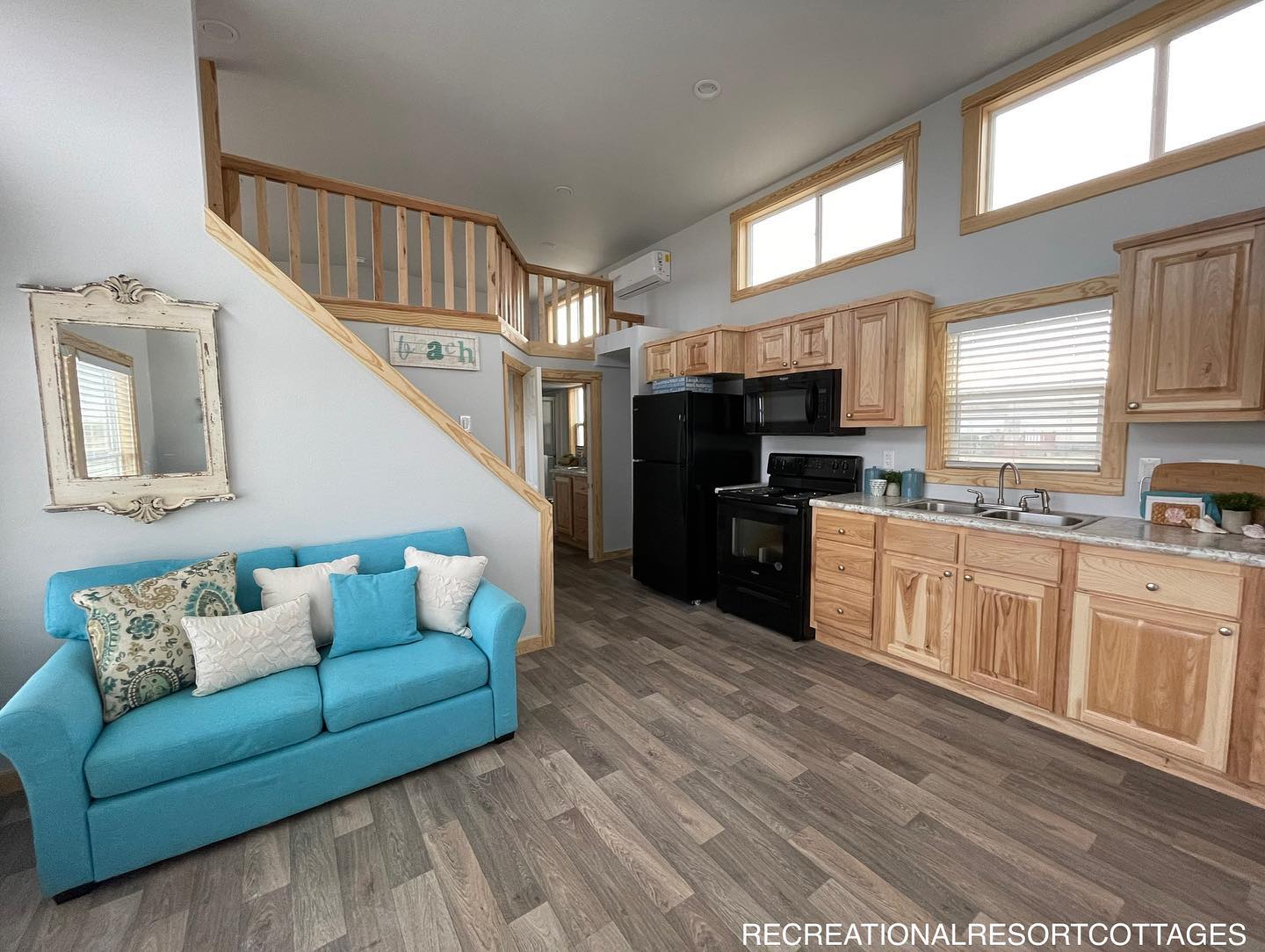 Bright, cozy tiny home interior with wooden cabinets and a staircase. A turquoise sofa with pillows, mirror, and large windows create a welcoming feel.