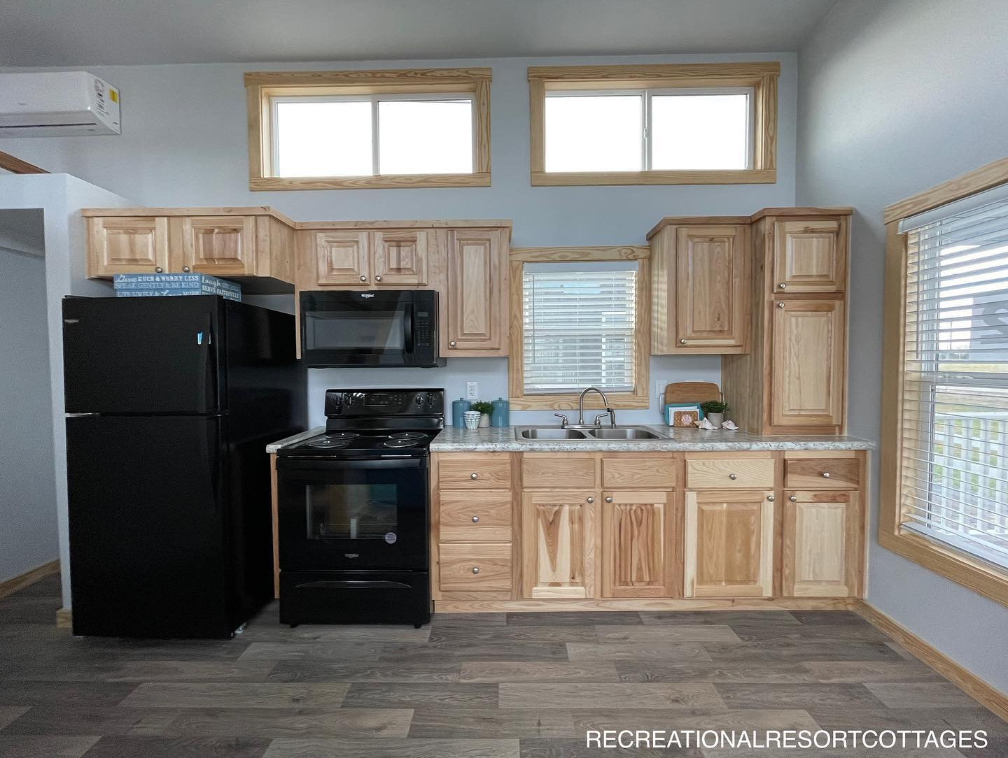 Compact kitchen with light wood cabinetry, black appliances, and a countertop sink beneath a window. Bright, modern, and neatly organized space.