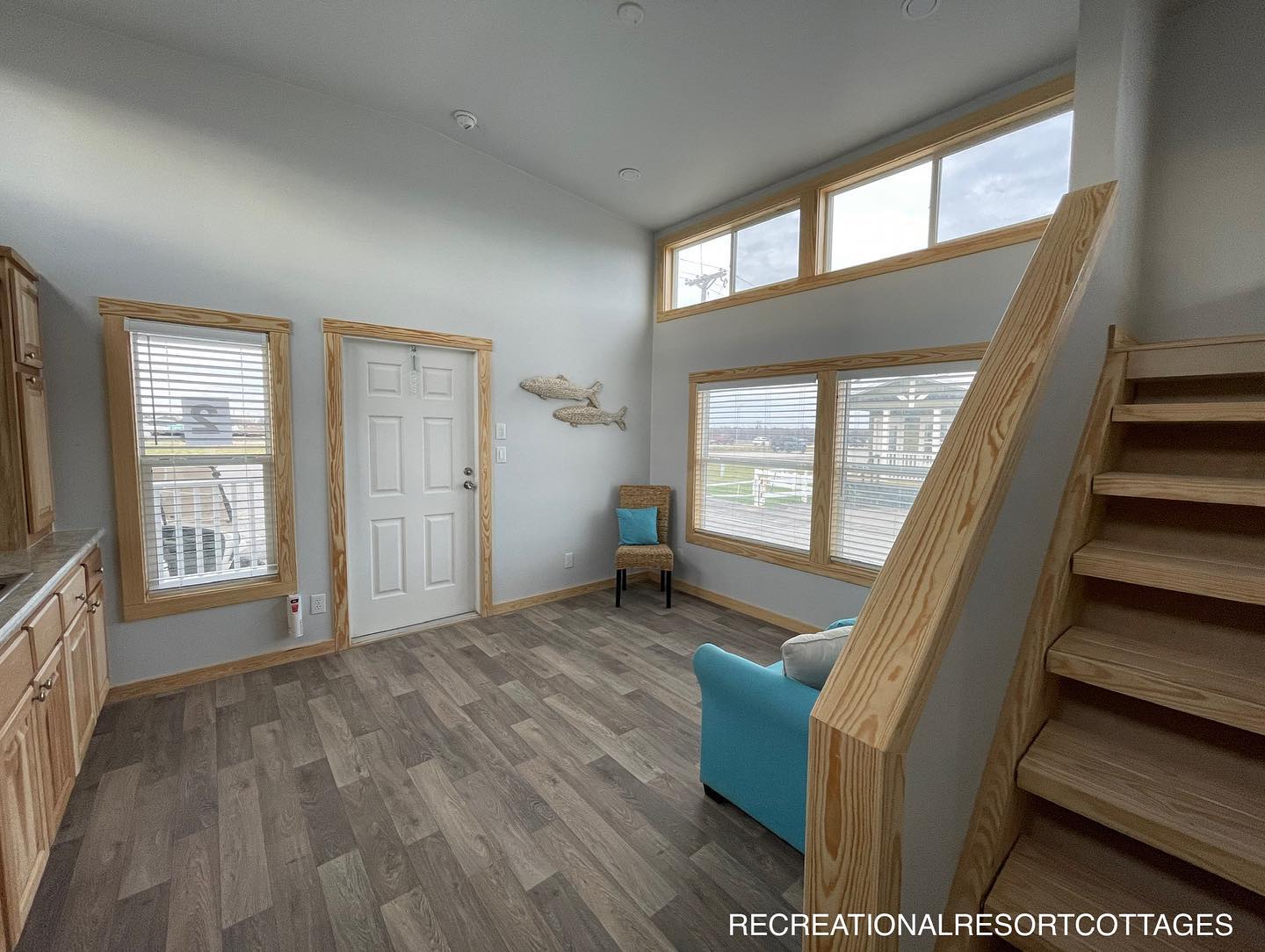 Bright, cozy room with high ceilings, wooden staircase, and large windows. Features a blue armchair, wicker chair, and fish wall art, creating a serene atmosphere.