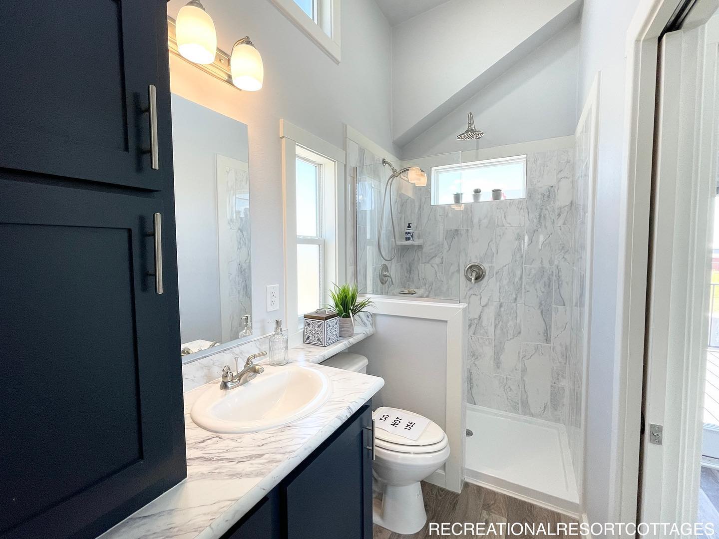 Bright bathroom with marble tile shower, white sink, and dark cabinets. Features large mirror, potted plant, and high ceiling with natural light.
