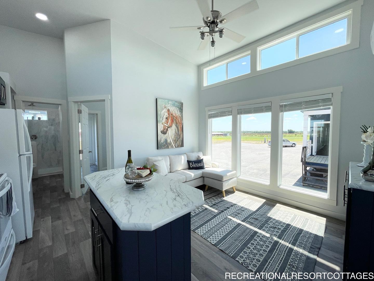 Bright living space with large windows, a white corner sofa, and a horse painting. Marble countertop island, ceiling fan, and a patterned rug add elegance.