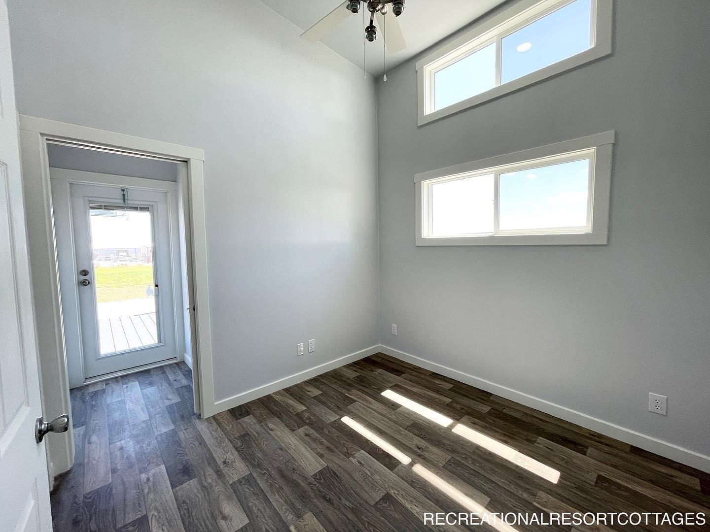 Alt text: "Bright, minimalistic room with high ceiling and light gray walls. Two slim rectangular windows let in natural light. Dark wood flooring and a door with a window lead to an outside area. A modern ceiling fan adds a contemporary touch."
