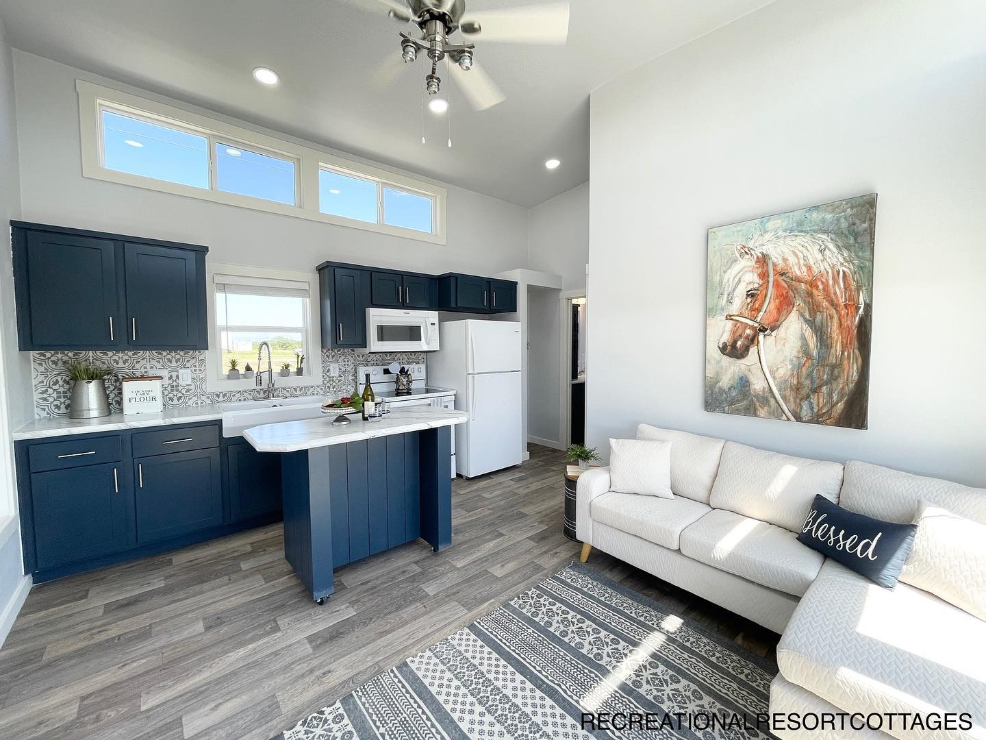 Bright, modern living area with a kitchen featuring blue cabinets, white counters, and appliances. A cozy sofa with a "Blessed" pillow, horse artwork, and patterned rug enhance the space.