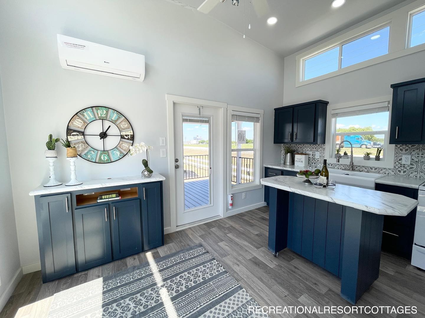 Modern kitchen with navy cabinets, marble countertops, and an island. A large clock adorns the wall, and natural light fills the space through windows and a glass door.