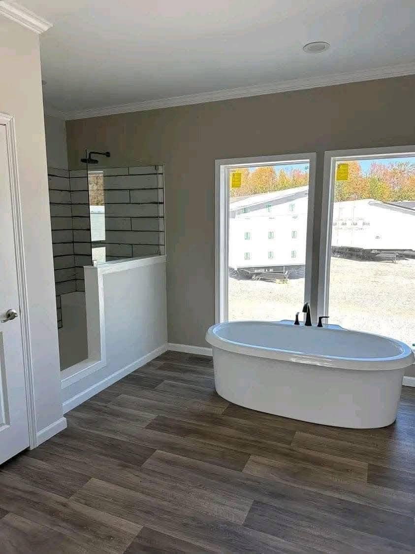 A modern bathroom with wood flooring, featuring a freestanding oval bathtub beside two large windows and a walk-in shower with subway tiles, creating a serene ambiance.