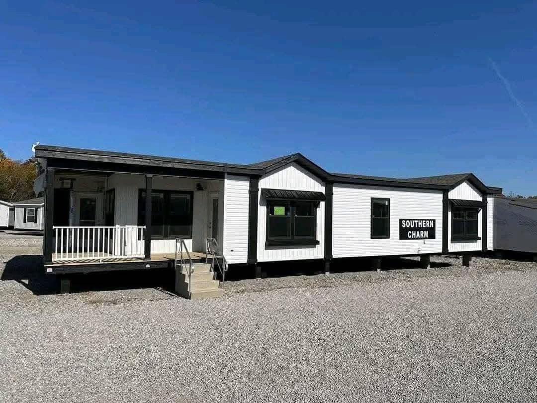 A modern, white mobile home with "Southern Charm" on the side, sits on gravel under a clear blue sky. It features a small porch with steps.
