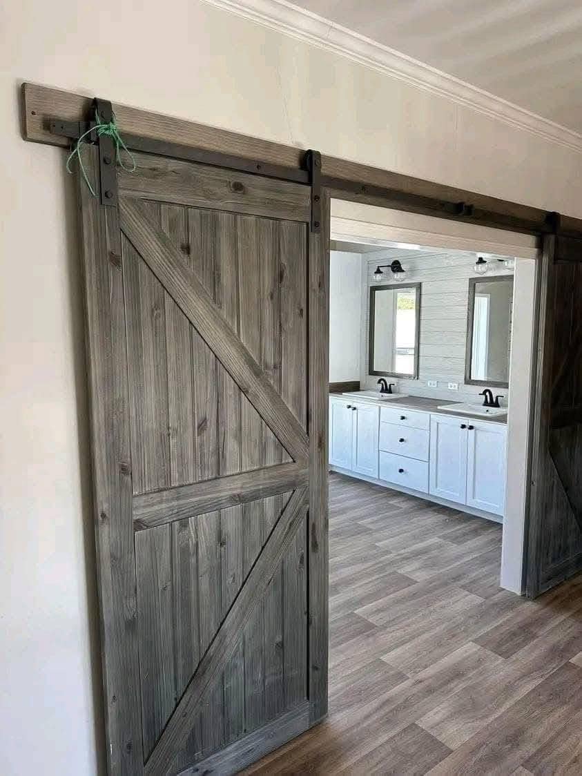 Rustic wooden barn door on black metal tracks slides open, revealing a modern bathroom with white cabinets, dual sinks, and warm wood flooring.