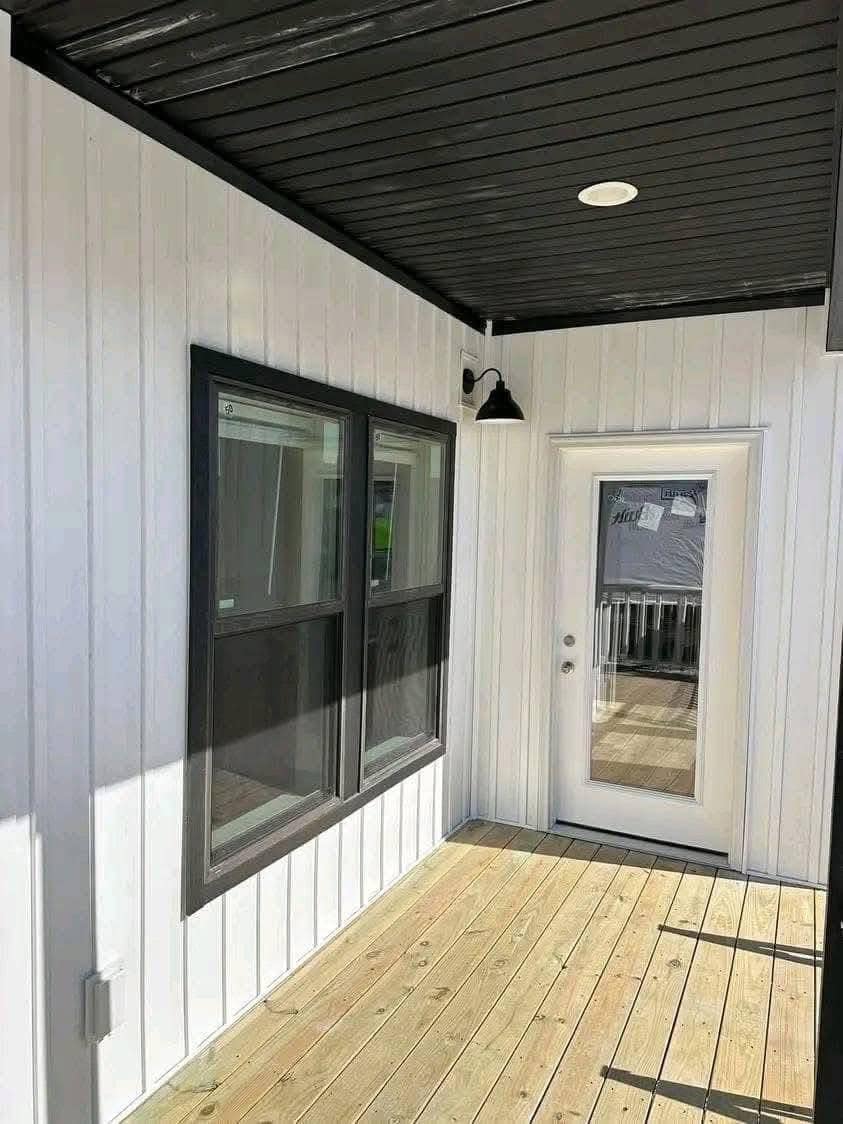 A covered porch features white siding, two dark-framed windows, a glass door, and a black ceiling with a light. The wood floor enhances the clean, modern look.