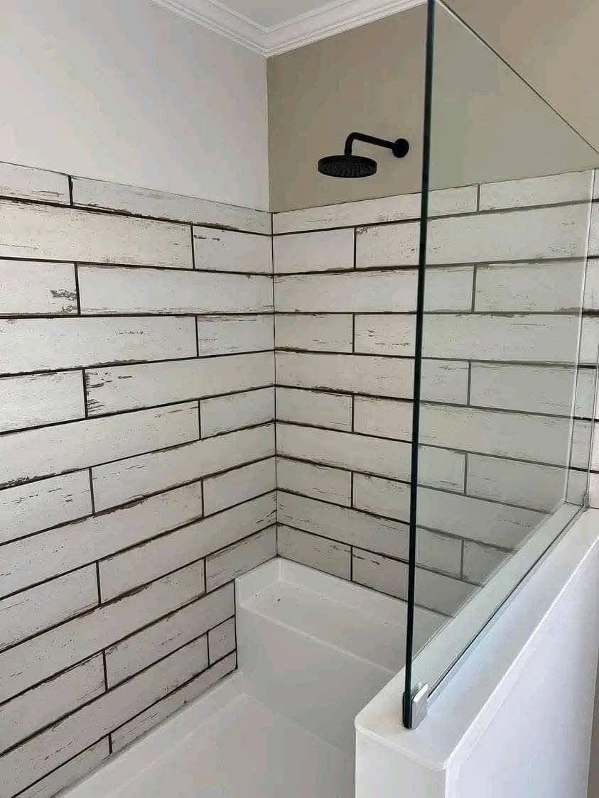 Corner of a bathroom with white subway tiles featuring distressed texture. Glass partition and black rainfall showerhead add modern elements.