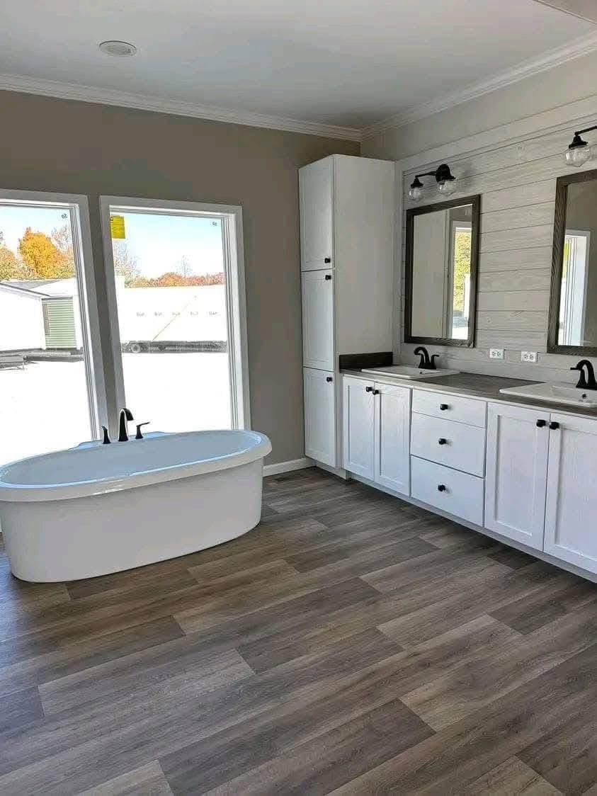 A modern bathroom with a freestanding white bathtub near large windows, double vanity with two mirrors, matte black fixtures, and wood-patterned flooring.