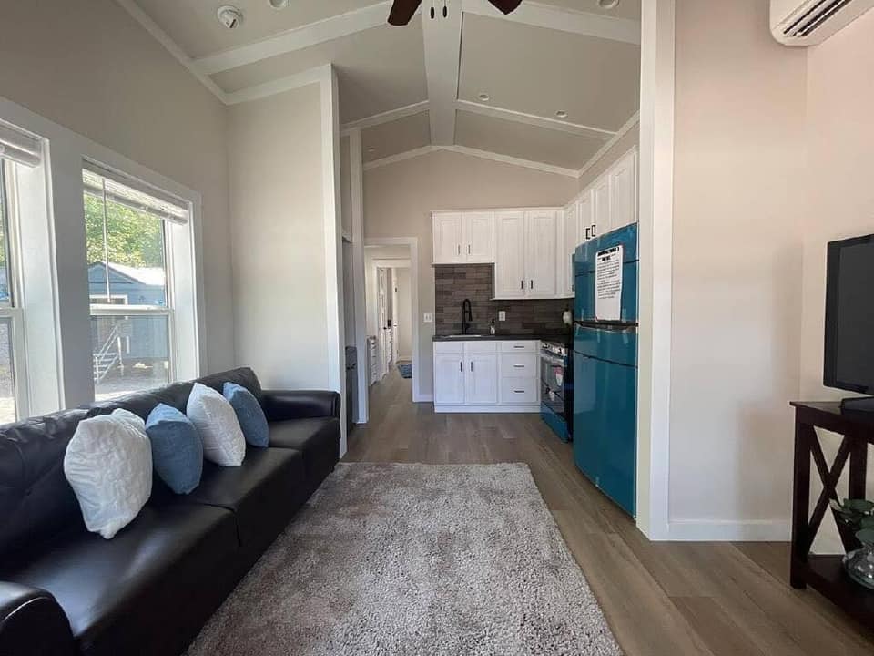 Modern living room with a black leather sofa, white and blue pillows, and a light rug. Bright kitchen with teal appliances and white cabinets. Cozy and bright ambiance.