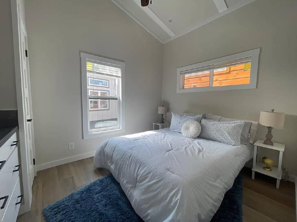 A cozy bedroom with soft natural light. It features a neatly made bed with white and gray bedding, two lamps on side tables, and a plush blue rug. The room has light wooden floors and white walls, creating a serene and welcoming atmosphere.