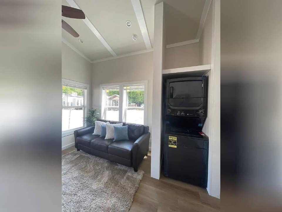 A cozy living room with a dark leather sofa and three assorted throw pillows. Large windows illuminate the space. A stacked washer-dryer is visible in a nook.