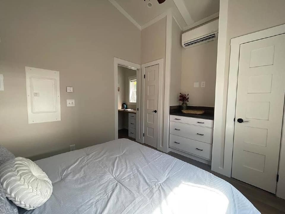 A cozy bedroom features a neatly made bed with a white comforter. There’s a dresser with a vase of flowers, two closed doors, and an air conditioner above. Soft, neutral tones create a peaceful ambiance.