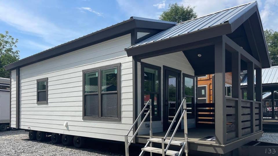 A modern tiny house with a white exterior and dark trim under a clear blue sky. It features a small porch with steps and a metal roof, evoking simplicity and coziness.