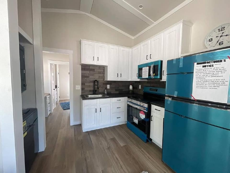 Compact kitchen with white cabinets and teal appliances, including a fridge and oven. Light wood flooring, a wall clock, and a brick-style backsplash. Bright and modern.