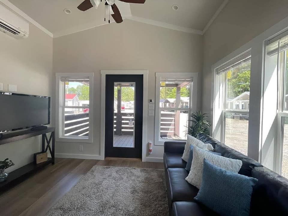 Modern living room with light walls and a high ceiling, featuring a ceiling fan, large windows, a dark sofa with blue and white pillows, and a TV on a stand. A door opens to a wooden deck, adding an inviting feel.