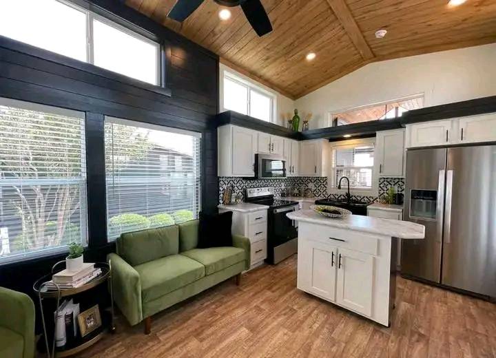 Modern kitchen and living area with wood ceiling, large windows, and a light-filled ambiance. Features include green sofas, a central island, and stainless steel appliances.