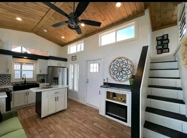 Modern tiny home interior with high wooden ceiling, ceiling fan, and natural light. Compact white kitchen, sleek appliances, and black accents. Cozy staircase leads upstairs.