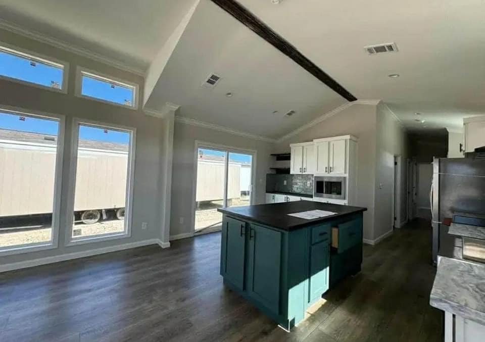 Open kitchen with large windows, providing natural light. Features a teal island, dark countertops, white cabinets, and dark wood flooring. Airy and modern.