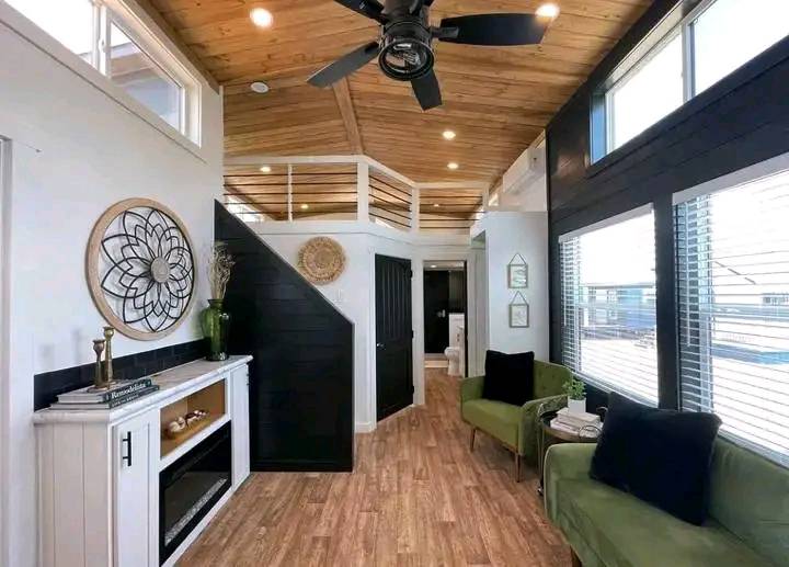 Cozy modern living room with wooden ceiling and floor, black fan, green armchairs, wall art, large windows, and natural light creating a warm ambiance.