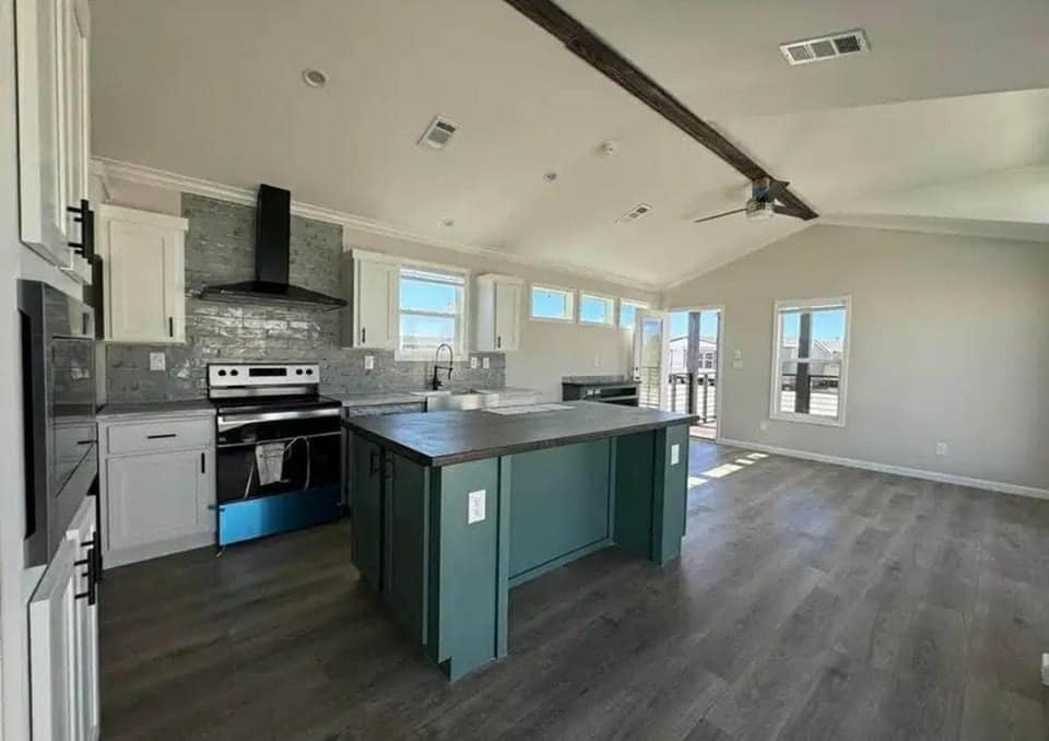 Modern kitchen with vaulted ceiling, wooden beams, and ceiling fan. Features a large teal island, stainless steel appliances, and light wood flooring.
