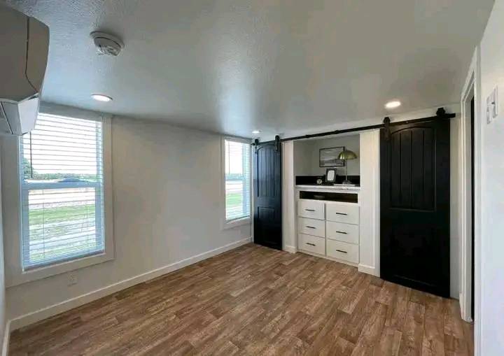 Minimalist room with wood flooring, white walls, recessed lighting, two large windows, and a dark sliding barn door revealing a dresser and mirror.