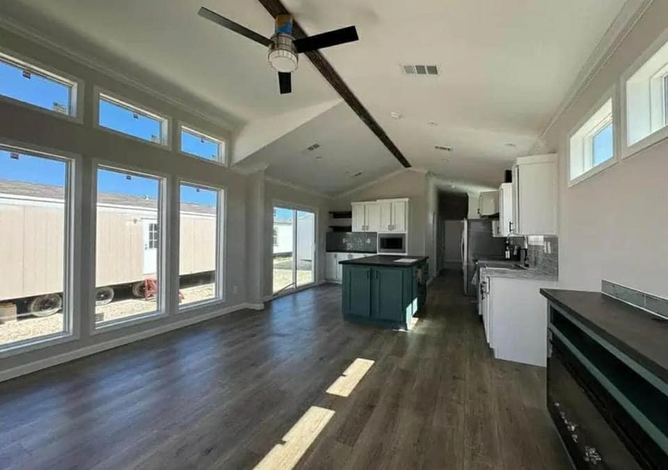 Bright, modern kitchen and living area with large windows, wooden floors, and a ceiling fan. The space has white cabinets and a dark green island.