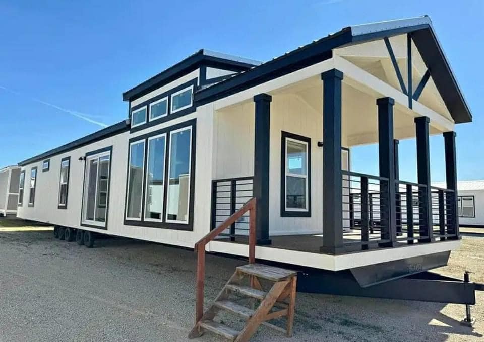Modern mobile home with large windows and a covered porch, featuring black and white accents, set on gravel under a clear blue sky.