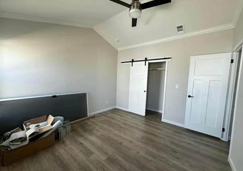 A minimalist room with light wood flooring, beige walls, and a ceiling fan. It features a white sliding barn door closet. A box and materials are in the corner, suggesting ongoing setup.
