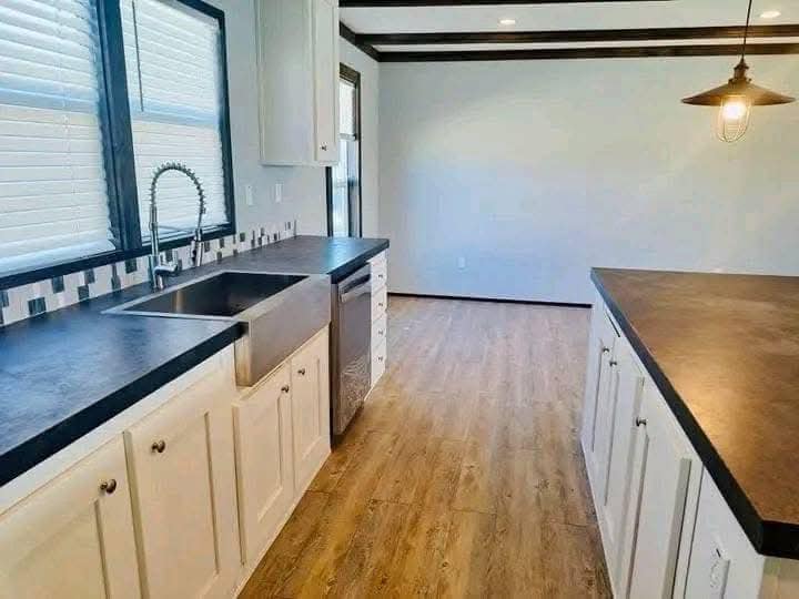 Modern kitchen with white cabinets, black countertops, and farmhouse sink. Wood flooring, overhead pendant light, and natural light from windows. Warm and inviting.