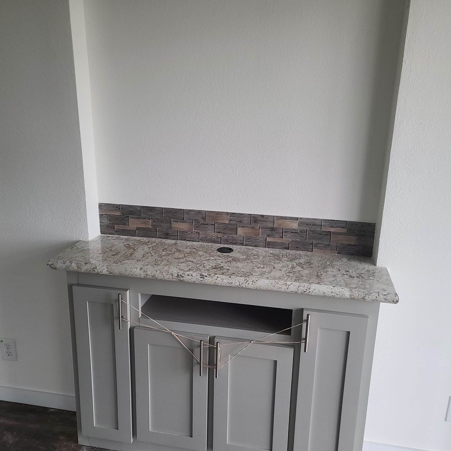 Gray cabinet with tied handles below a speckled granite countertop. A tiled backsplash with a hole is set against a plain white wall.