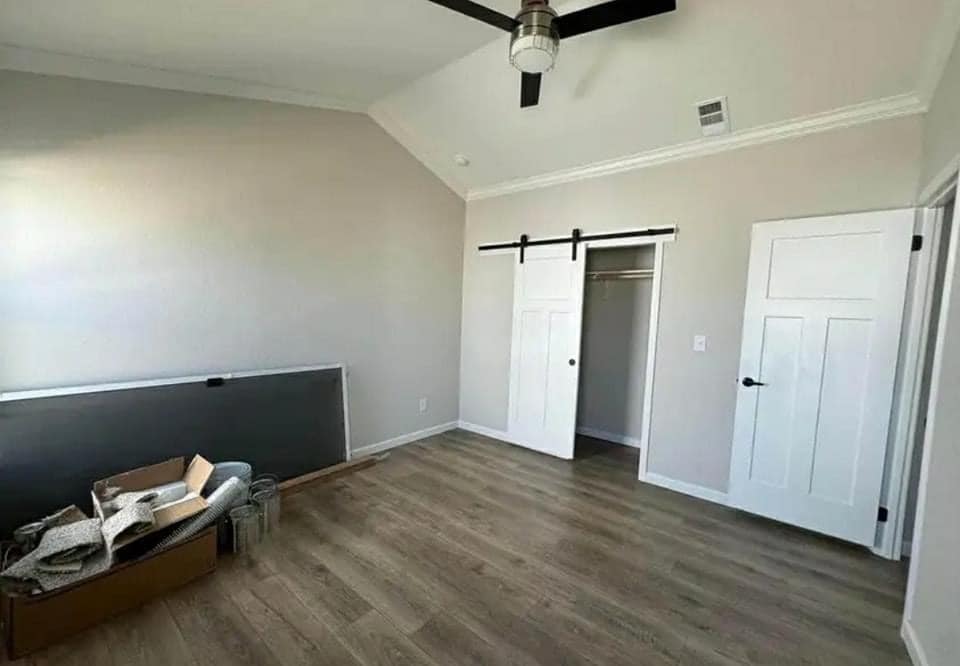 Spacious room with light wood floors, pale walls, and a ceiling fan. A sliding barn door reveals a closet. Boxes and fabric rest on the floor, suggesting recent moving.
