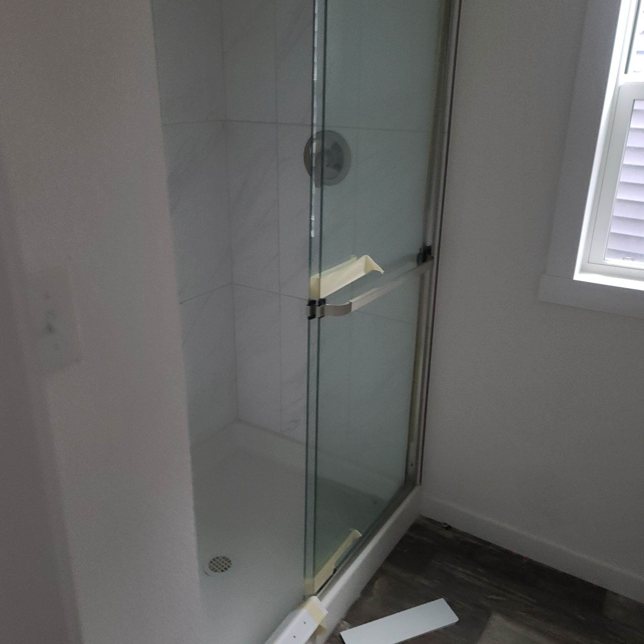 A small bathroom with a glass shower enclosure. The shower has a sliding door with a towel bar and white tiled walls. Natural light from a window brightens the space.