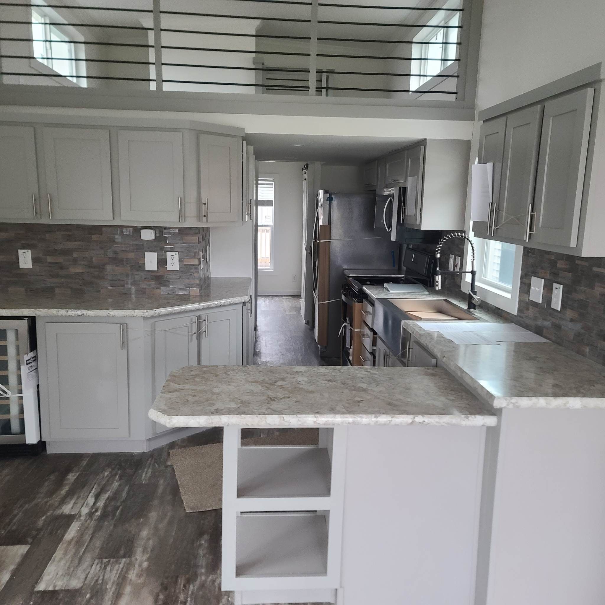 Modern kitchen with light grey cabinets, stainless steel appliances, and a stone-patterned backsplash. A loft area overlooks the space, enhancing openness.