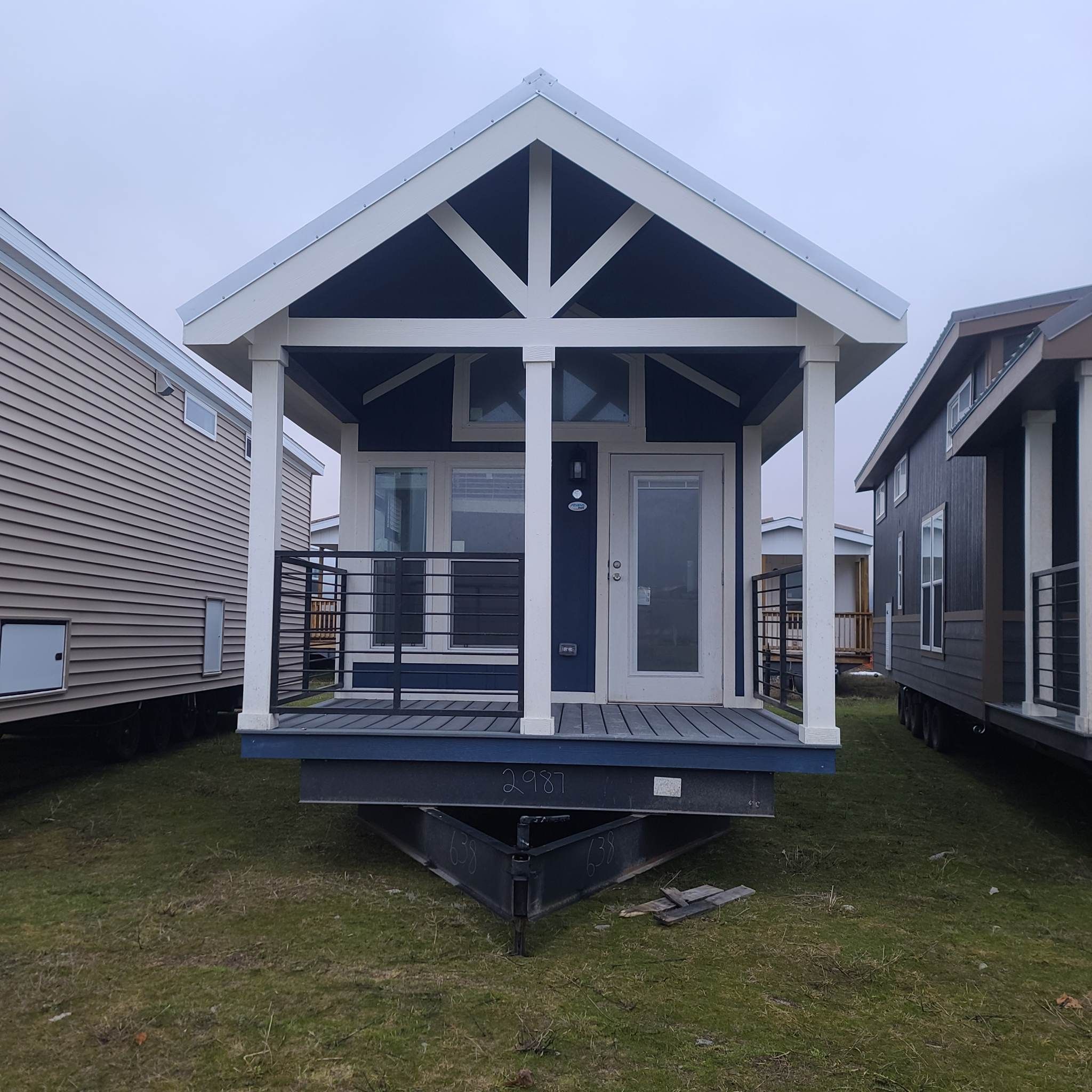 A small, modern navy and white tiny house with a front porch sits on a grassy area between other similar homes under a cloudy sky.